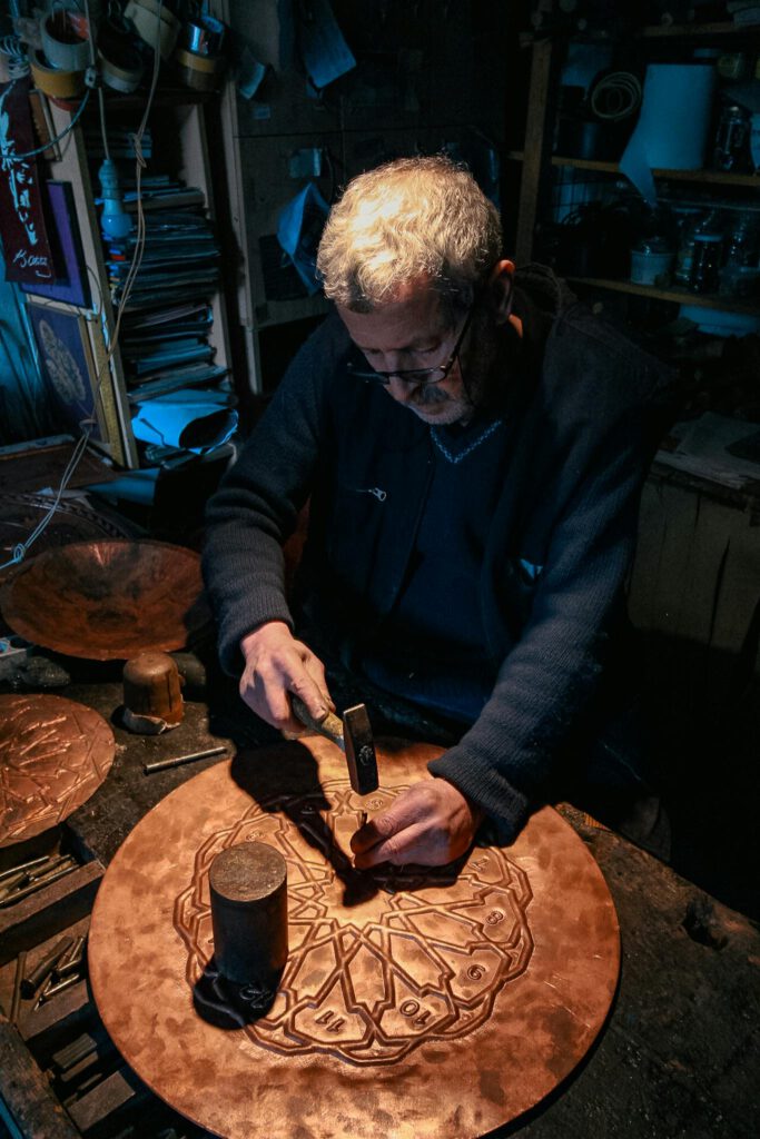 An artisan working intently on a decorative copper plate in a cozy workshop setting.
